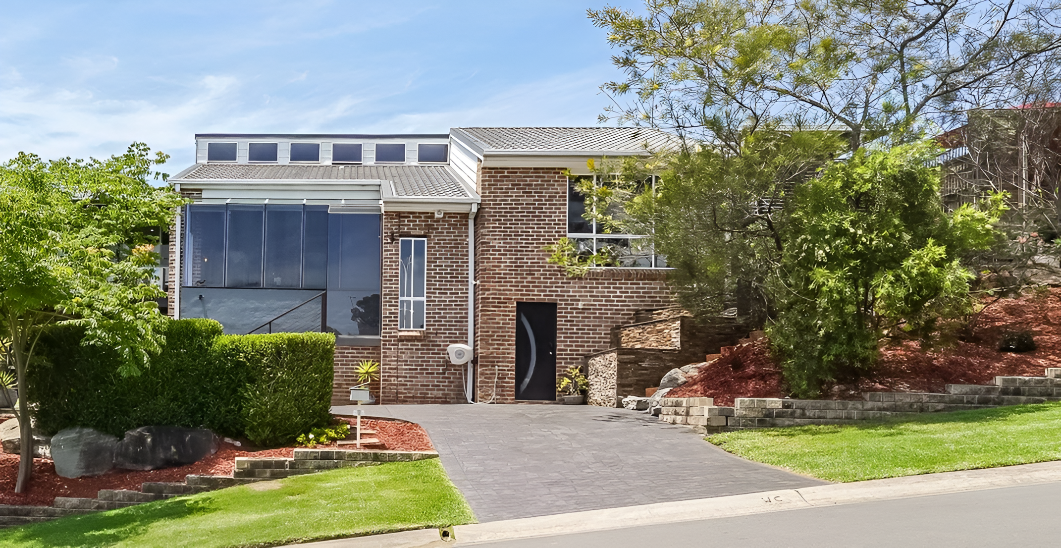 Modern house with brick exterior, large glass windows, a chimney, and landscaped front yard with trees, shrubs, and a driveway.